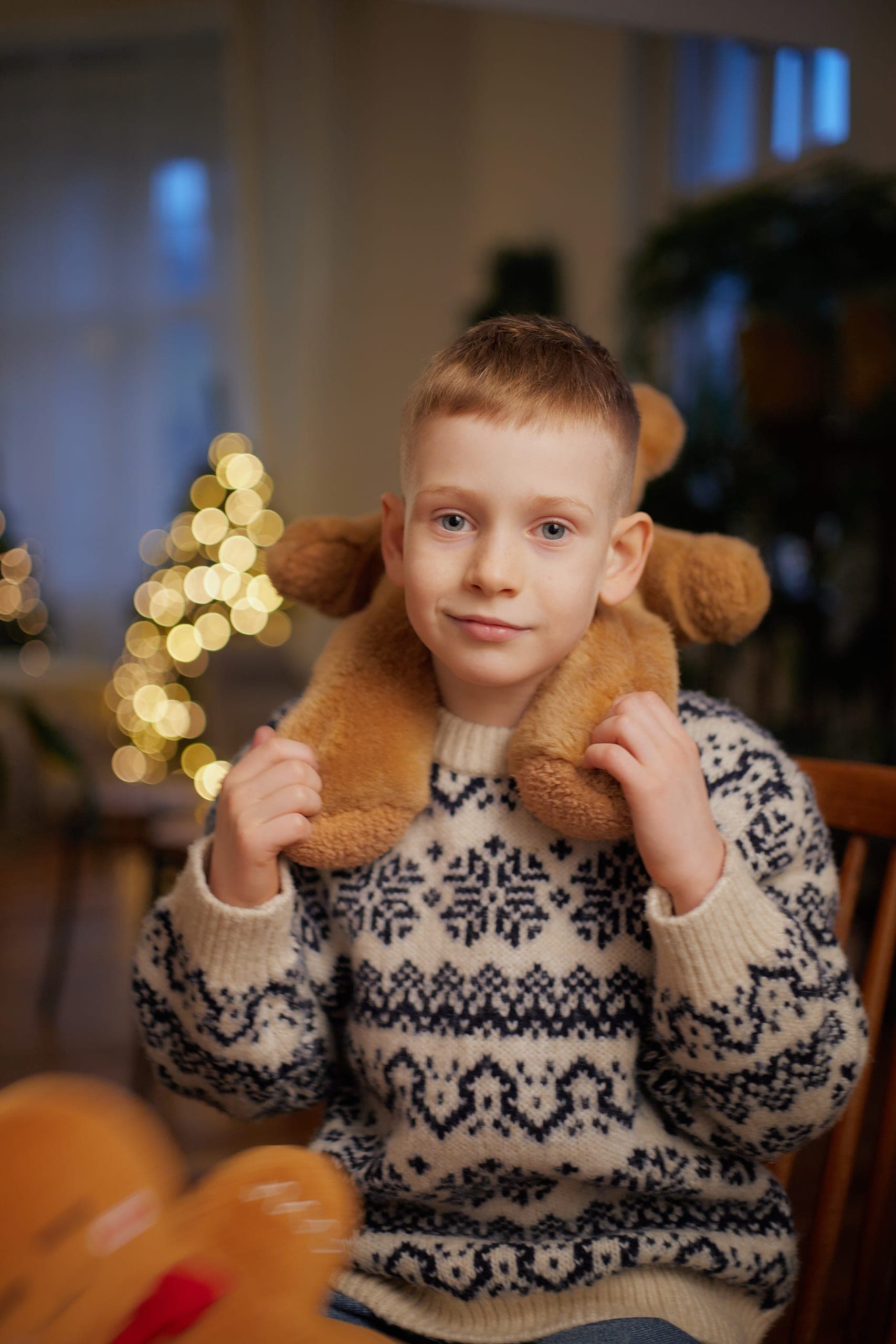 Chłopiec w świątecznym swetrze z pluszowym misiem na ramionach, w tle światełka choinki, sesja rodzinna
