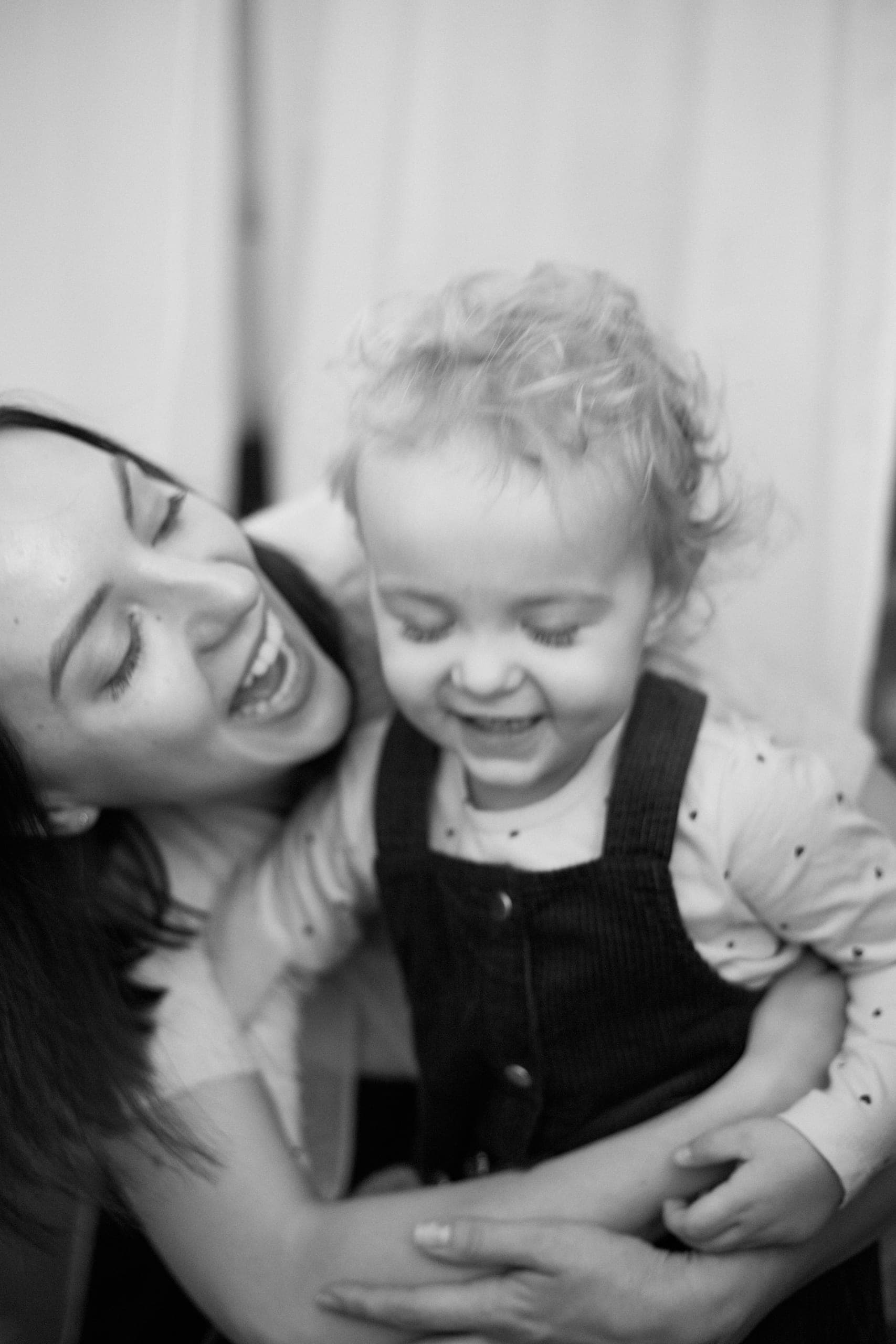 Roześmiana mama tuli kręconowłosą córeczkę w ogrodniczkach – czarno-biały portret z sesji rodzinnej