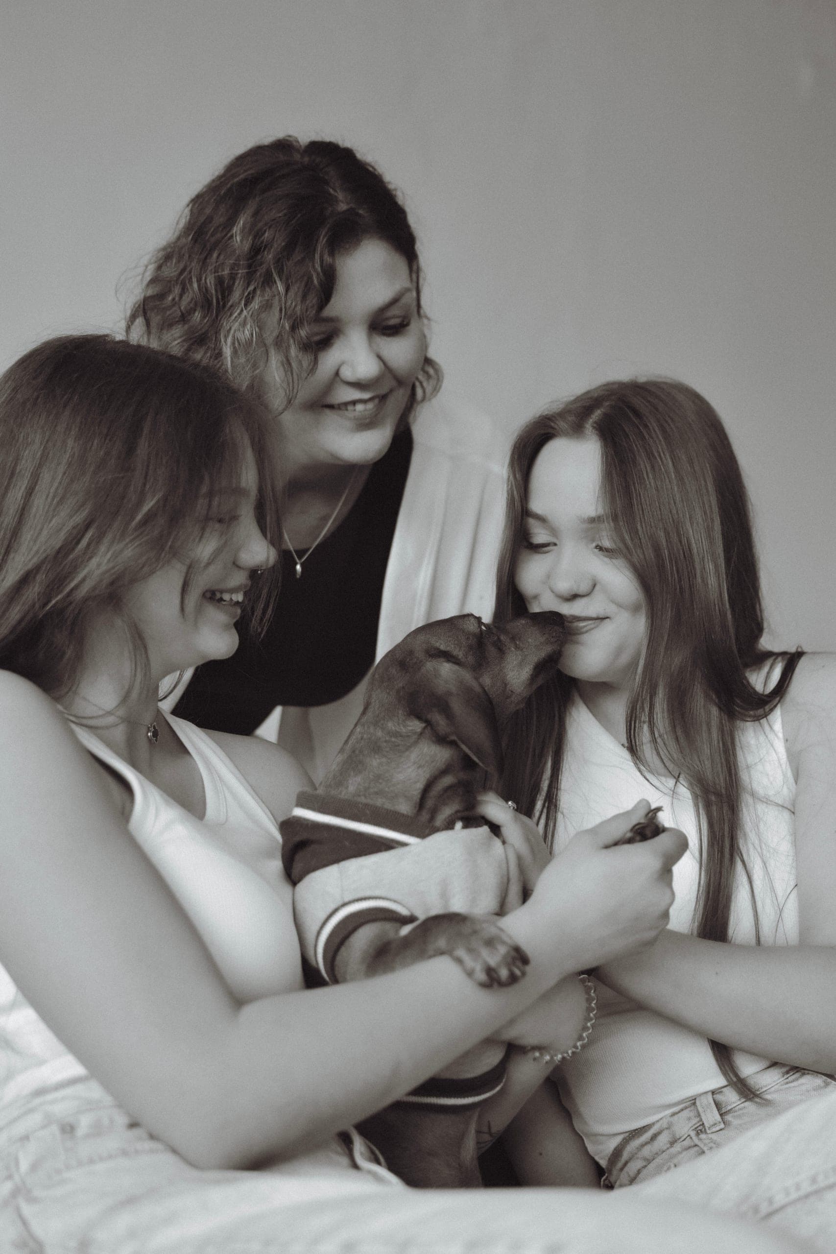 Mama z dwiema córkami śmieje się, gdy szczeniak jamnika liże twarz dziewczyny, czarno-biała sesja rodzinna