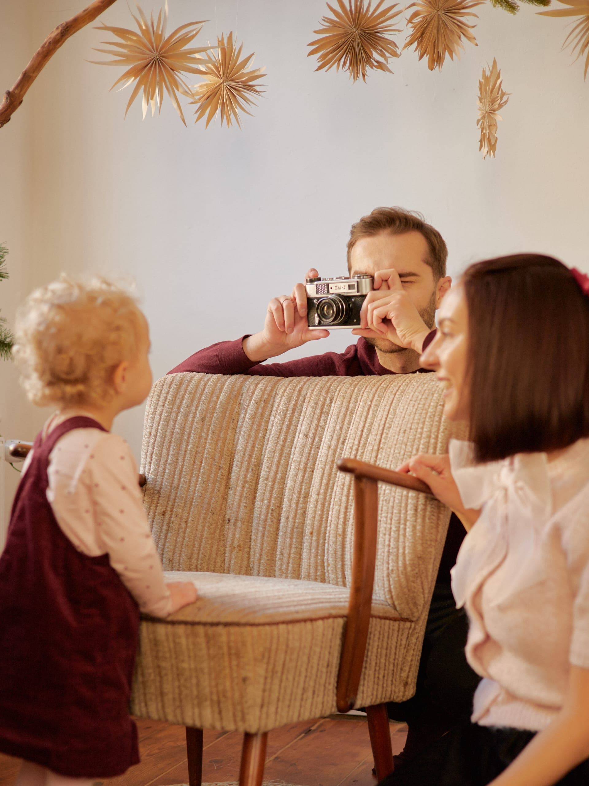 Tata fotografuje córeczkę starym aparatem zza fotela, mama obok – świąteczna domowa sesja rodzinna