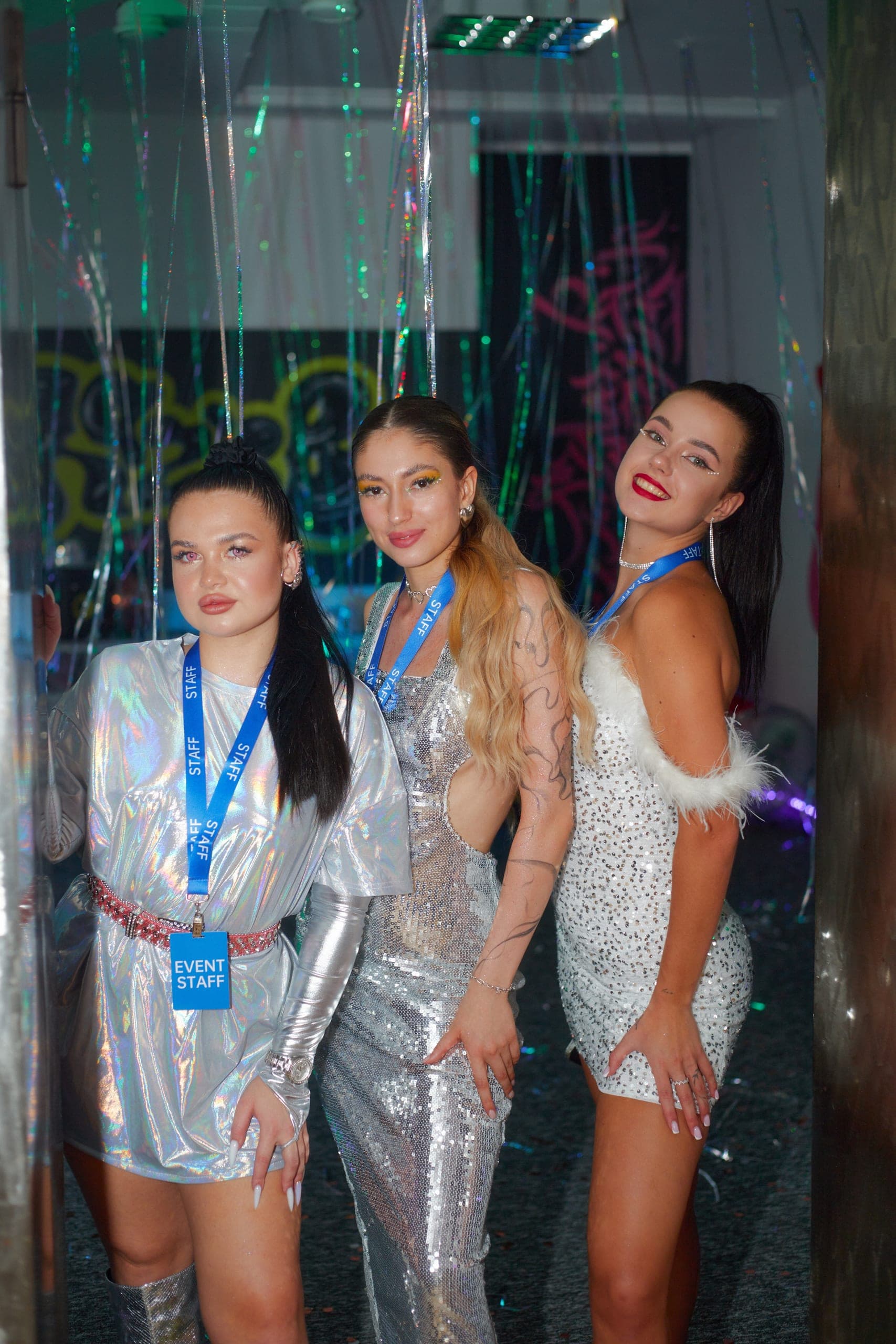 Trzy kobiety w błyszczących srebrnych sukienkach pozują wśród lametty na imprezie – fotografia reportażowa z eventu