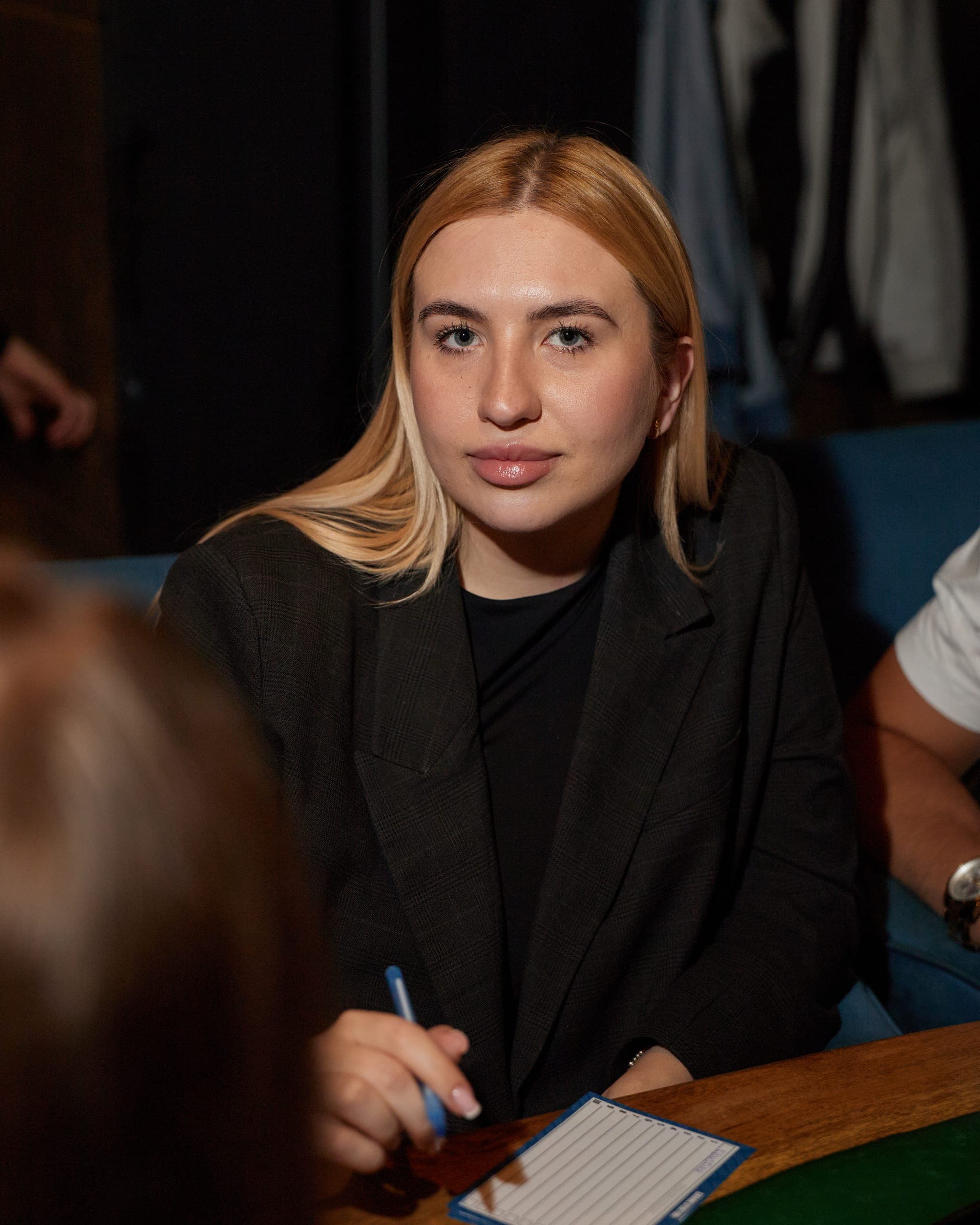 Blondynka w czarnej marynarce siedzi przy stole z kartą odpowiedzi i patrzy w obiektyw podczas quizu